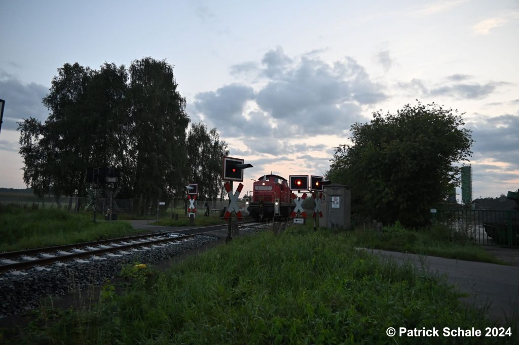 Diesellokomotive der Baureihe 294 erreicht bei Sonnenaufgang den Bahnhof Barenburg und passiert den Bahnübergang Birkenkampsweg mit eingeschalteten Blinklichtern.