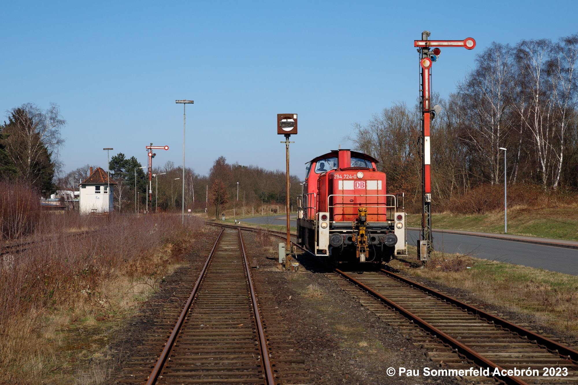 Diesellok 294 724 der DB Cargo AG in verkehrsrot ranigert im nordlichen Bahnhofskopf des Sulinger Bahnhofs und passiert dabei ein Gleissperrsignal und ein Hauptsignal; im Hintergrund befindet sich ein weiteres Hauptsignal und das Stellwerk Sulingen Nord (Sn)