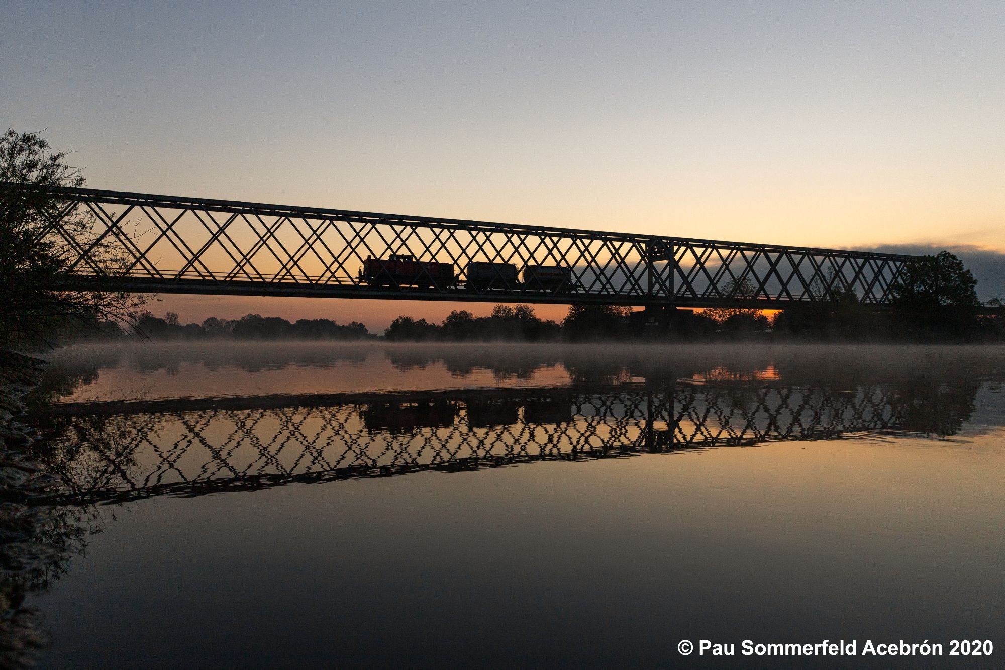 Diesellok 296 029 der DB Cargo AG passiert mit einem Güterzug aus 2 Wagen die Weserbrücke bei Nienburg (Weser). Der Zug spiegelt sich in der Weser und es hängt leichter Dunst über dem Wasser.