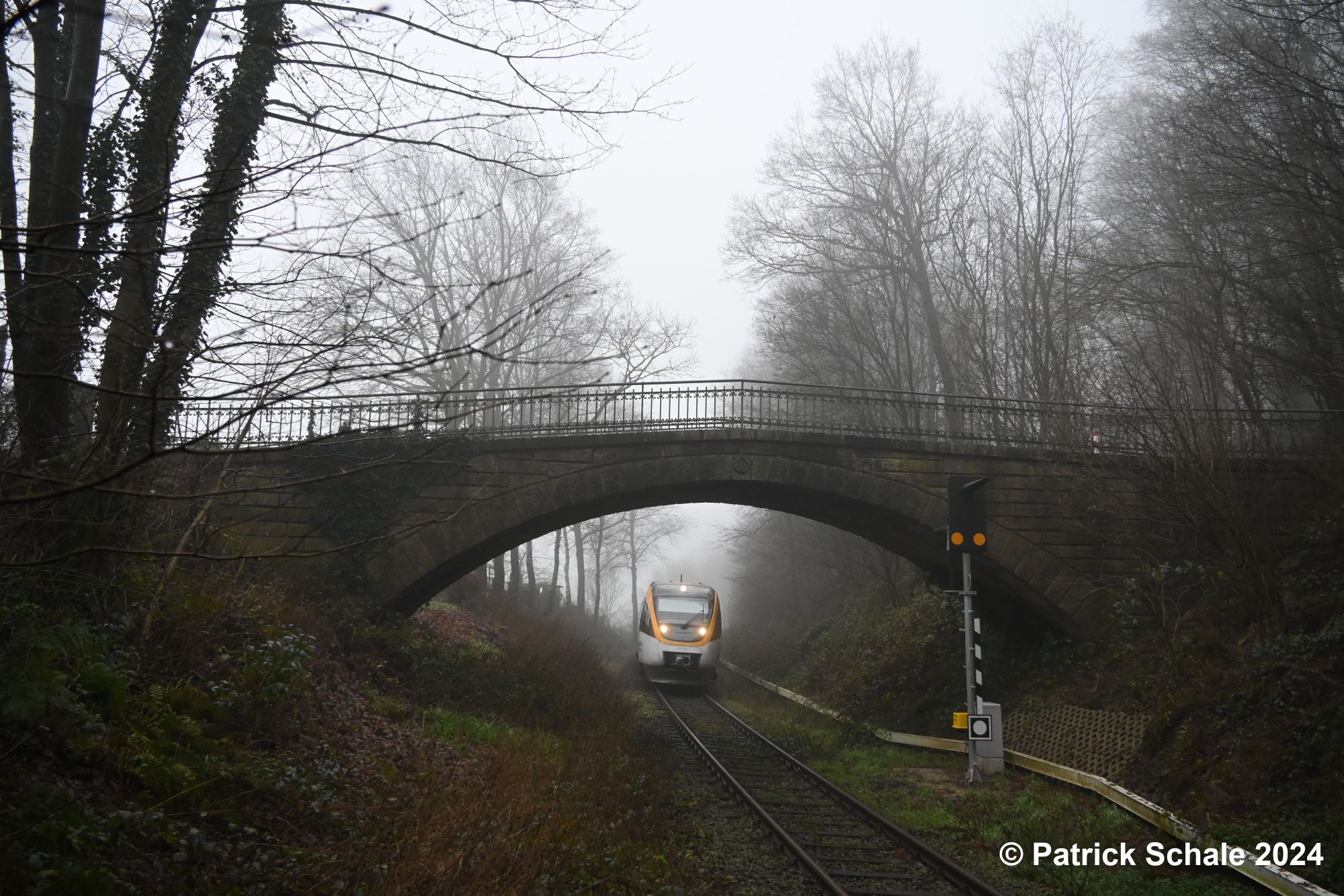 Dieseltriebwagen der Eurobahn der Linie RB 71 nach Bielefeld Hbf fährt an einem kalten und nebeligen Wintertag in den Haltepunkt Mesch Neue Mühle ein und passiert dabei eine Brücke aus Naturstein; rechts vor der Brücke steht ein Bahnüberwachungssignal