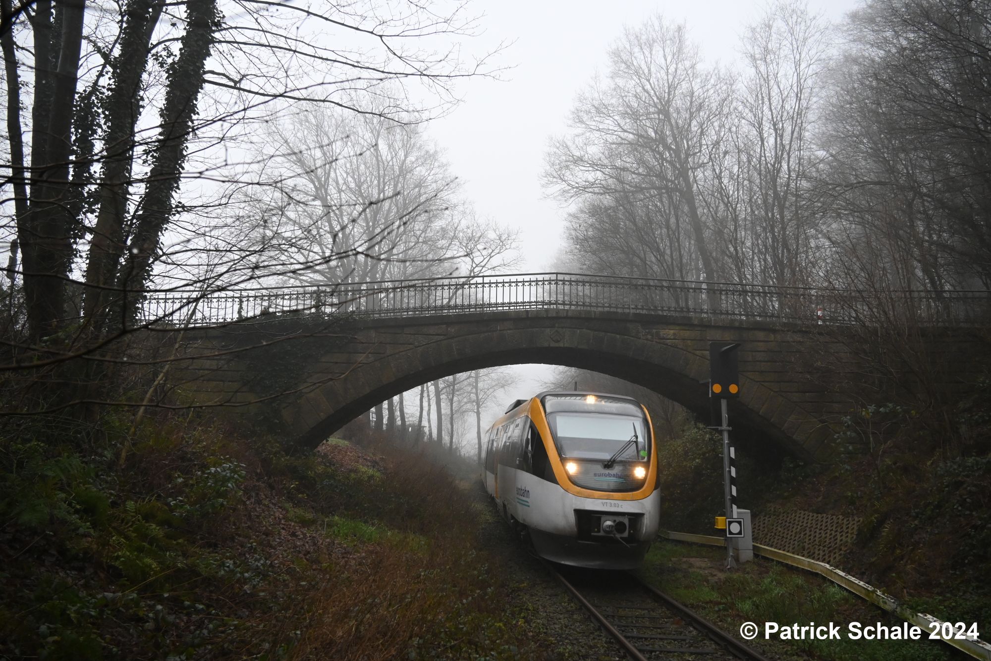 Dieseltriebwagen der Eurobahn der Linie RB 71 nach Bielefeld Hbf fährt an einem kalten und nebeligen Wintertag in den Haltepunkt Mesch Neue Mühle ein und passiert dabei eine Brücke aus Naturstein; rechts vor der Brücke steht ein Bahnüberwachungssignal