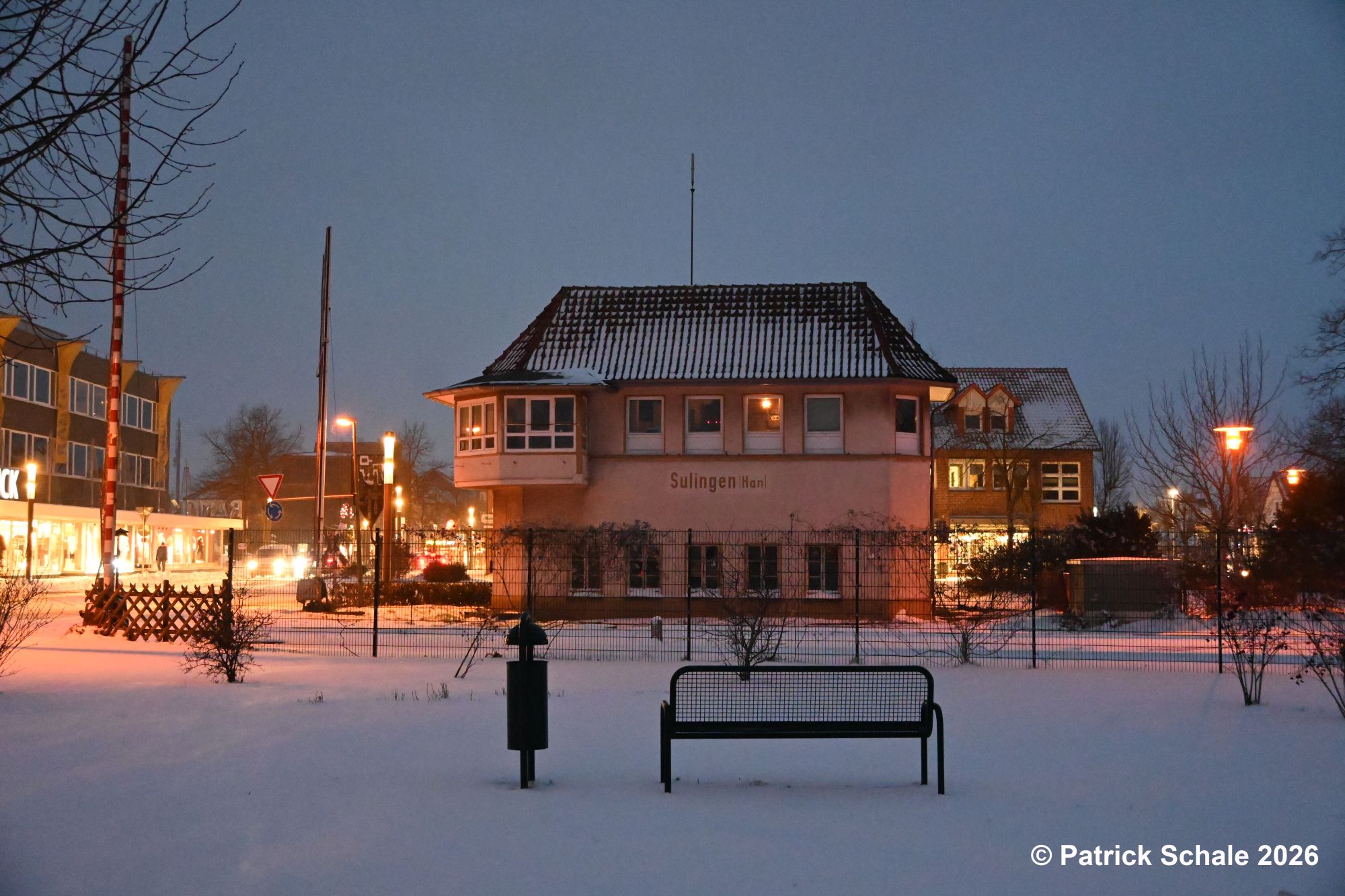 Stellwerk Sf im Schnee, im Vordergrund eine Sitzgelegenheit mit Mülleimer, links 2 Schrankenbäume vom Bahnübergang Nienburger Straße