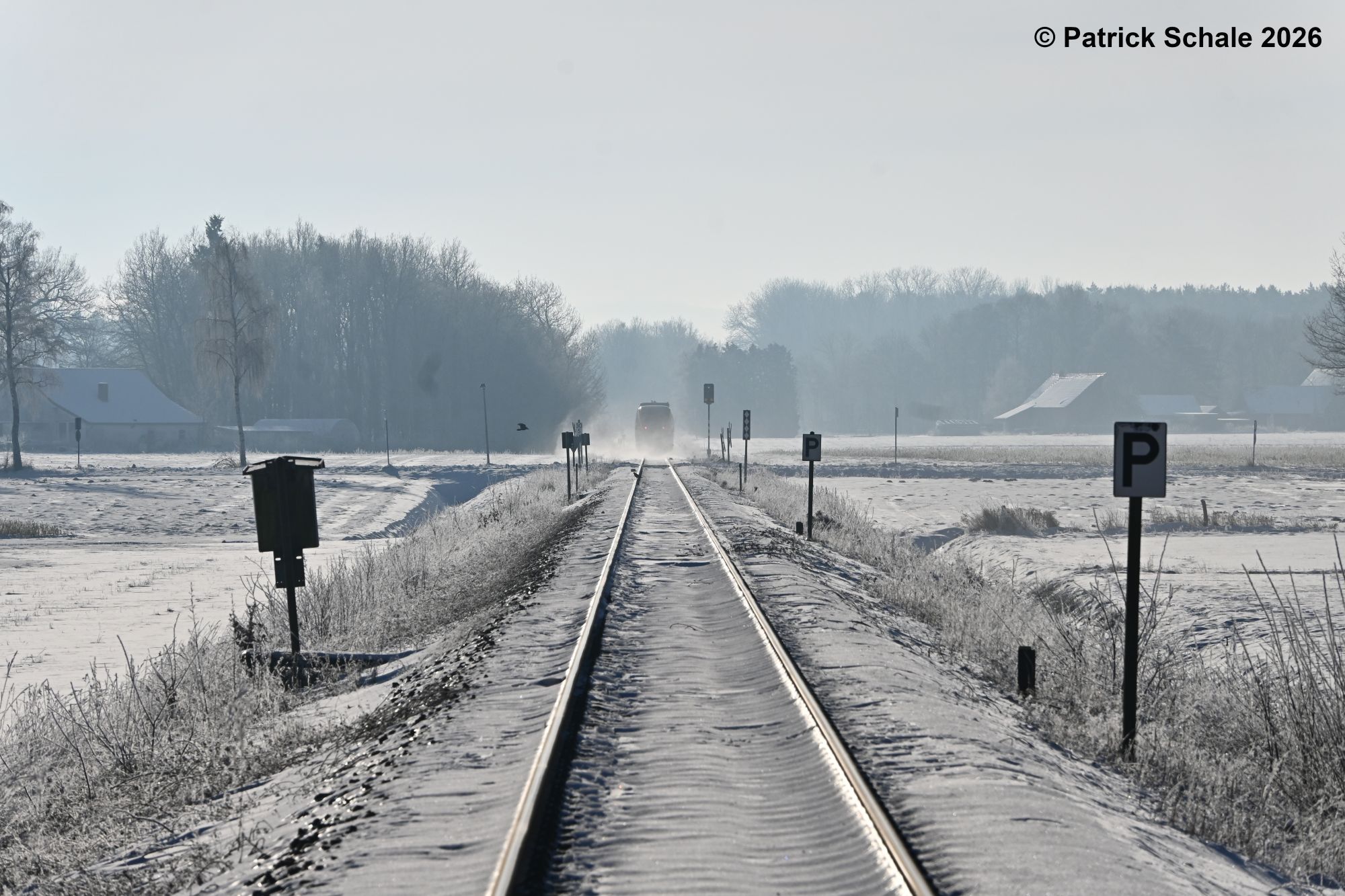 Triebwagen der Eurobahn als RB 71 nach Bielefeld Hbf hat einen nicht-technisch gesicherten Bahnübergang passiert und verschwindet hinter aufgewirbeltem Schnee, vorne P-Tafeln zur Absicherung des Bahnübergangs und links die Rückseite eines Fernsprechkastens