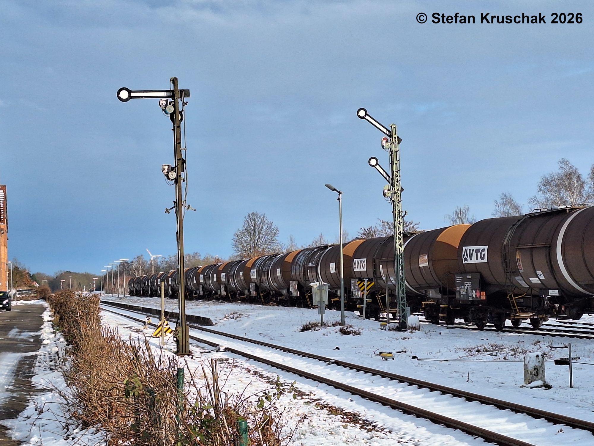 Kesselwagen, welche an der Diesellokomotive 261 029-3 der DB Cargo AG hängen, bei der Ausfahrt aus dem verschneiten Bahnhof Sulingen; Das rechte Ausfahrsignal zeigt HP 2 (Langsamfahrt).