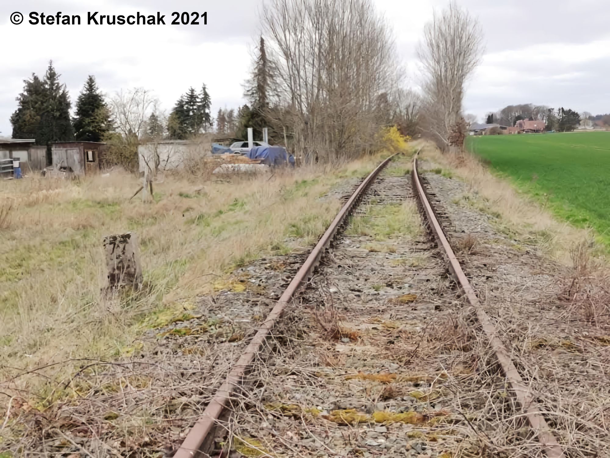 Stillgelegtes Gleis bei Kilometer 77,3, links vom Gleis stehen Schuppen, im Hintergrund Wohnhäuser