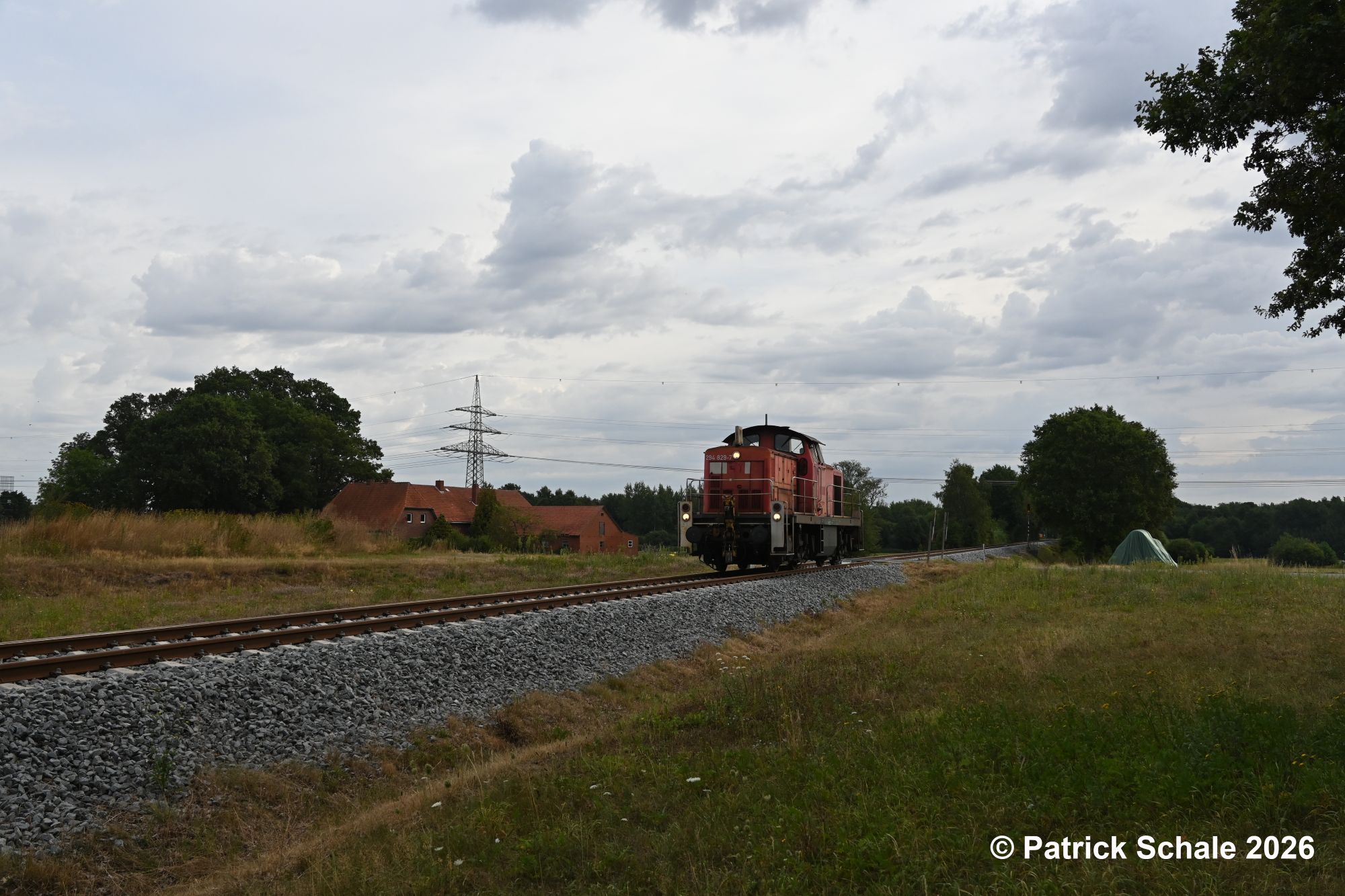 Diesellokomotive der Baureihe 294 in verkehrsrot passiert einen landwirtschaftlichen Betrieb auf dem Weg nach Diepholz im Bereich Sulingen-Vorwerk; im Hintergrund verläuft eine Hochspannungsleitung