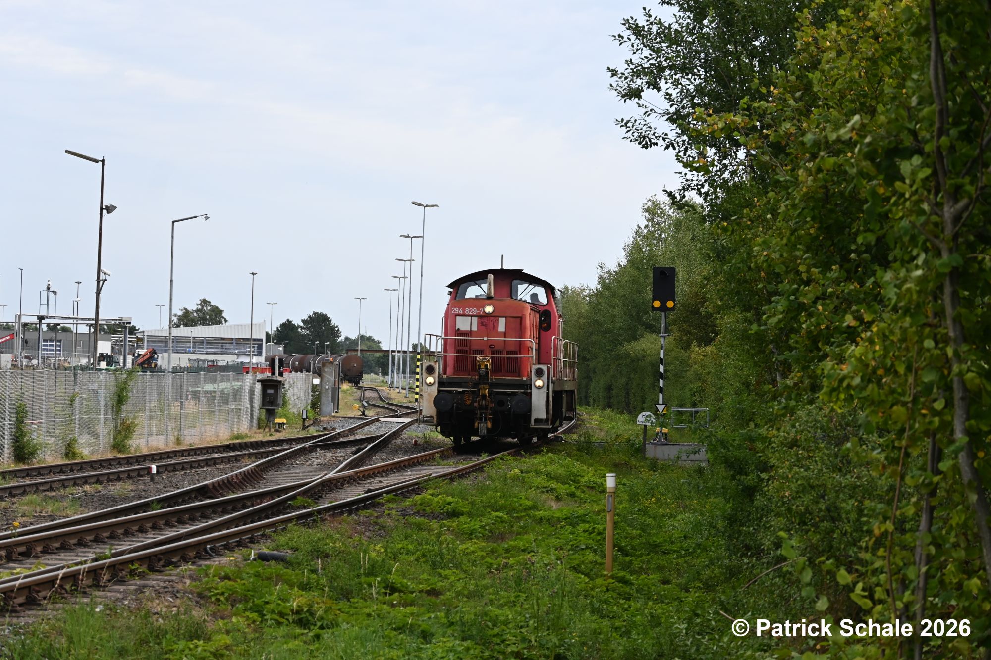 Diesellokomotive 294 829-7 ist in den Bahnhof Barenburg eingefahren und passiert gerade den Gleisanschluss zum Werk von ExxonMobil, wo die Lokomotive mit rohölbeladene Kesselwagen abholen wird. Rechts von der Lokomotive steht ein Bahnübergangüberwachungssignal.
