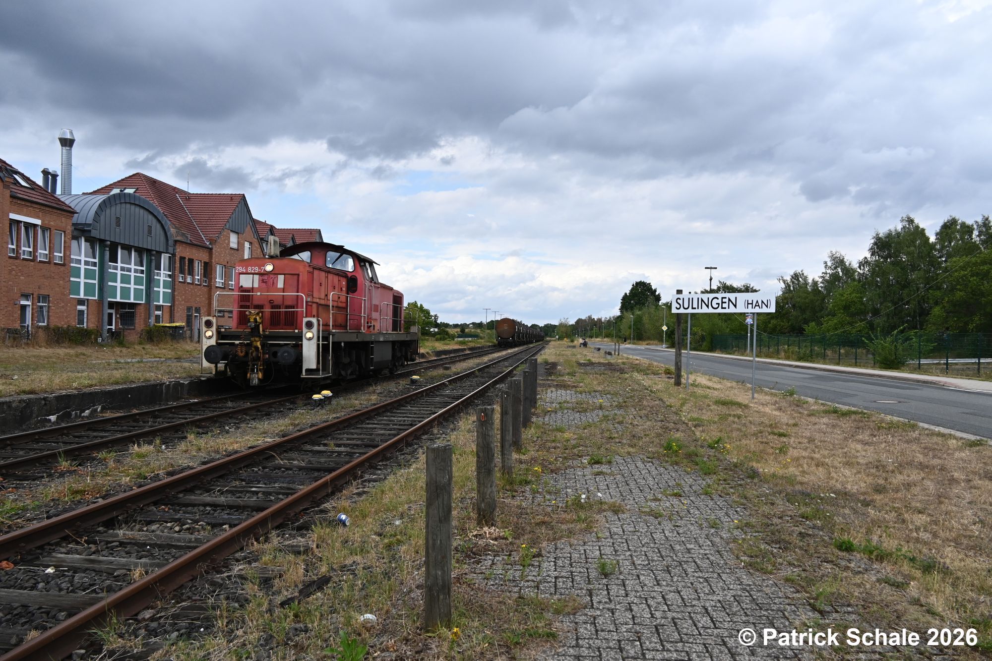 Diesellkokomotive der Baureihe 294 hat den im Hintergrund stehenden Kesselwagenpark über Gleis 4 umfahren und setzt sich nun zur Weiterfahrt wieder an die Kesselwagen. Rechts steht das Stationsschild und links ist ein Wohn- und Geschäftshaus zu sehen.